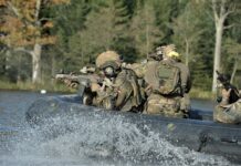 Drei Soldaten schießen bei der Übungsserie Taurus 2024 mit dem Gewehr aus einem schwarzen fahrenden Schlauchboot auf einem See.
